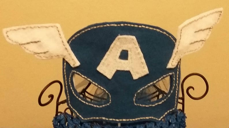 Op de afbeelding: Een blauw vilten masker met witte vleugels en een witte "A" in het midden. Het masker heeft oogopeningen en is afgewerkt met witte stiksels. De vleugels zijn wit met grijze stiksels. Het masker wordt op een decoratieve standaard getoond.