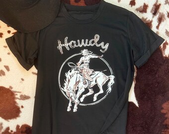 May include: Black t-shirt with a white graphic of a cowboy riding a bucking horse. The graphic includes the word "Howdy" in a rope design.