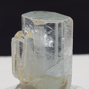 May include: A clear, light blue aquamarine crystal with a faceted, hexagonal shape. The crystal is sitting on a white, textured surface.