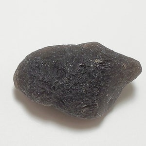 May include: A dark, irregularly shaped rock with a rough texture. Next to it is a black cube with the text "T." and "1 CM" on its sides. The rock appears to be a dark gray or brown color.