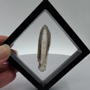 May include: A clear, smoky quartz crystal point in a black diamond-shaped display case.