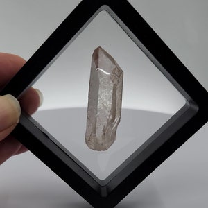May include: A clear quartz crystal point in a black diamond-shaped display case.