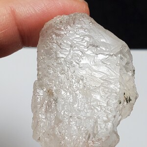 Può includere: Un cristallo trasparente, traslucido, con una texture ruvida e un aspetto leggermente nuvoloso. Il cristallo ha una forma unica e irregolare.