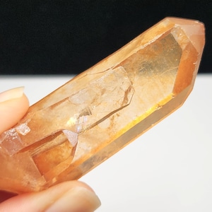 Könnte beinhalten: Ein durchscheinender, orangefarbener Kristall mit einer facettierten, spitzen Spitze. Der Kristall wird in einer Hand gehalten und zeigt seine natürliche, raue Textur und innere Muster. Die Farbe des Kristalls reicht von hell bis dunkelorange.