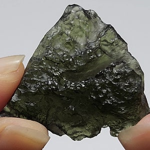 May include: A dark green, translucent, irregularly shaped stone with a rough, textured surface. The stone is being held between two fingers.