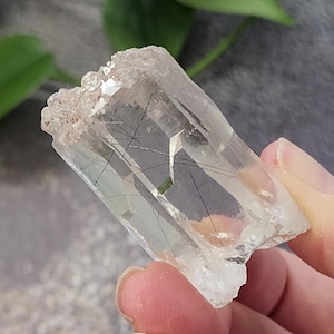 Puede incluir: Un cristal de cuarzo transparente con inclusiones negras. El cristal es puntiagudo en un extremo y tiene una superficie rugosa y natural.