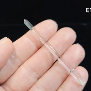 May include: A clear, slender crystal wand, held in a hand. The wand is transparent with a pointed tip and a rectangular base. The background is a solid black.