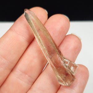 May include: A clear, elongated crystal with a light brown hue, held in a hand. The crystal has a pointed tip and a textured surface, catching the light. The background is a mix of black and white.