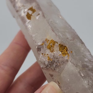 Puede incluir: Un cristal de cuarzo transparente con inclusiones doradas. El cristal tiene forma de prisma largo y delgado y una superficie lisa y pulida.