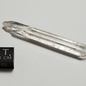 Puede incluir: Una punta de cristal de cuarzo transparente con un acabado rugoso y natural. El cristal mide aproximadamente 12,7 cm de largo y se muestra junto a un cubo de 1 cm para la escala.