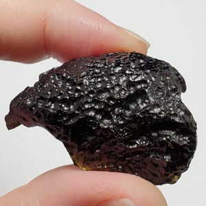 May include: A dark, black, and iridescent tektite with a rough, textured surface. The tektite has a unique, irregular shape.