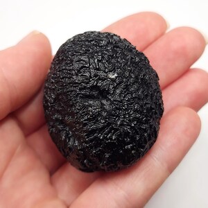 May include: A dark, textured, black stone held in a hand. The stone has a rough, uneven surface with a slightly rounded shape. The overall appearance suggests a natural, possibly geological specimen.