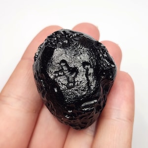 May include: A dark, glossy black tektite stone with a textured surface. The natural stone has an irregular shape and is held in a hand against a white background. The tektite appears to have a unique pattern on its surface.