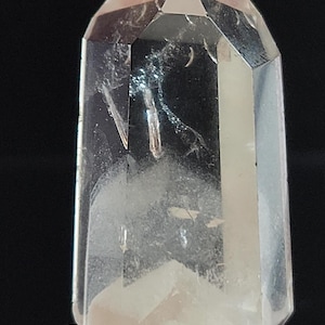 May include: A clear quartz crystal point with a hexagonal shape. The crystal is held between fingers against a black background. The quartz has internal inclusions and a cloudy appearance.