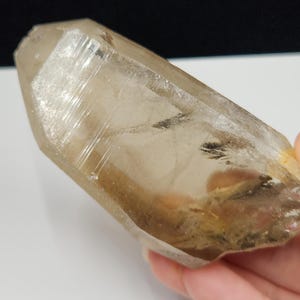 May include: A translucent, light brown smoky quartz crystal point with a hexagonal shape. The mineral specimen features visible internal inclusions, natural striations along its faceted surface, and a pointed termination with clear, glassy edges.