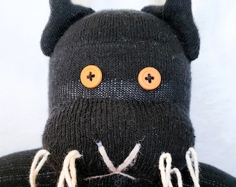 May include: A black sock monkey with orange button eyes and white yarn whiskers. The monkey has black sock horns and is made from a black knitted sock.