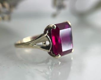 May include: A gold ring featuring a large, rectangular, deep red gemstone. The ring's setting has an ornate, openwork design on the sides, and the gemstone is the focal point. The ring is set against a soft, blurred background.