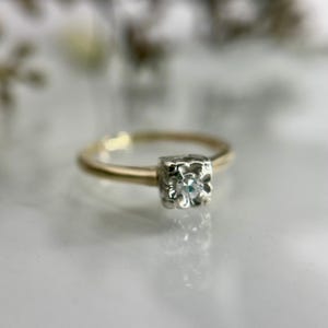 May include: A gold ring with a square, floral-patterned setting. The setting holds a small, round, clear diamond. The ring is set against a blurred, neutral background, highlighting the ring's details.