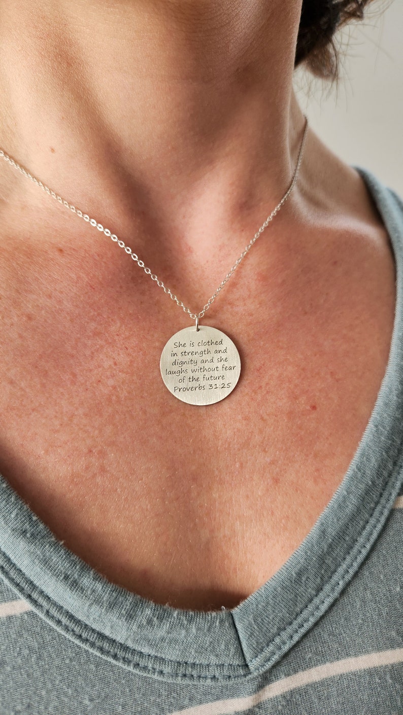 Model in teal and white striped shirt is wearing large round silver necklace with large round pendant, engraved on front with Scripture verse in 6 lines of text.