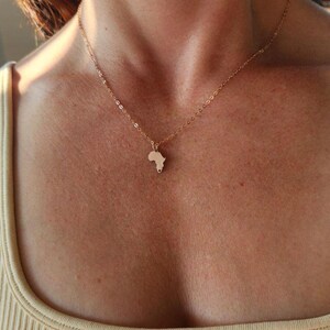 Three necklaces with Model in tan ribbed shirt is shown wearing a tiny rose gold Africa country outline necklace. There is a cutout heart in the very bottom of the charm on South Africa.