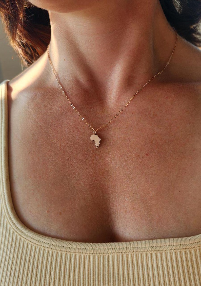 Three necklaces with Model in tan ribbed shirt is shown wearing a tiny rose gold Africa country outline necklace. There is a cutout heart in the very bottom of the charm on South Africa.