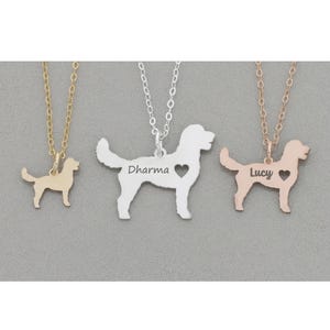 Three Goldendoodle dog outline necklaces in silver, gold, rose gold lay on dark gray stone background. Silver in middle with heart cutout and Dharma engraved, gold is tiny with no heart; rose gold is small on right side with Lucy engraved.