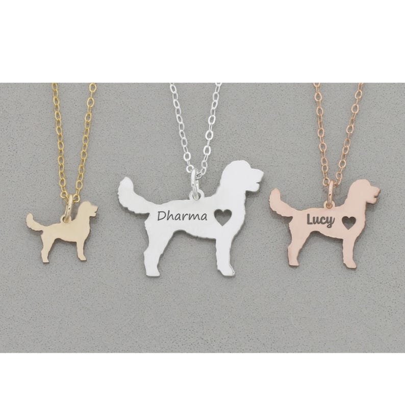 Three Goldendoodle dog outline necklaces in silver, gold, rose gold lay on dark gray stone background. Silver in middle with heart cutout and Dharma engraved, gold is tiny with no heart; rose gold is small on right side with Lucy engraved.
