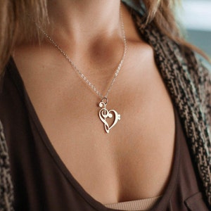 Pode incluir: Um colar prateado com um pingente em forma de cora&ccedil;&atilde;o com uma clave de sol e notas musicais. O pingente pende de uma corrente delicada. O colar &eacute; usado sobre uma blusa marrom.