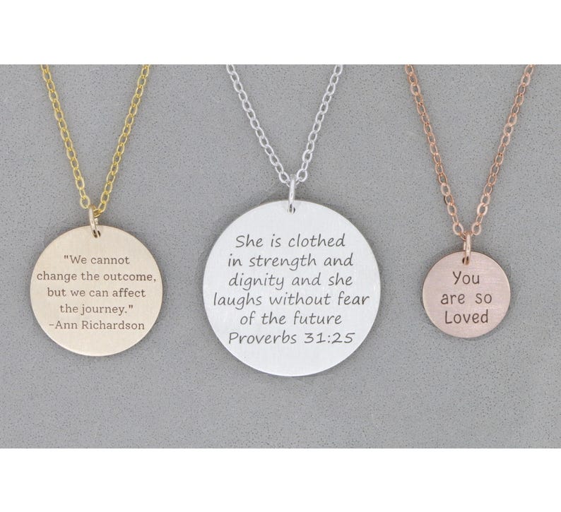 Three necklaces in gold, silver, and rose gold lay next to eachother on gray stone background. Each round pendant is engraved with different quotes in 3 different sizes.