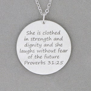 Silver necklace with large round pendant, engraved on front with Scripture verse in 6 lines of text is laying on gray stone background.
