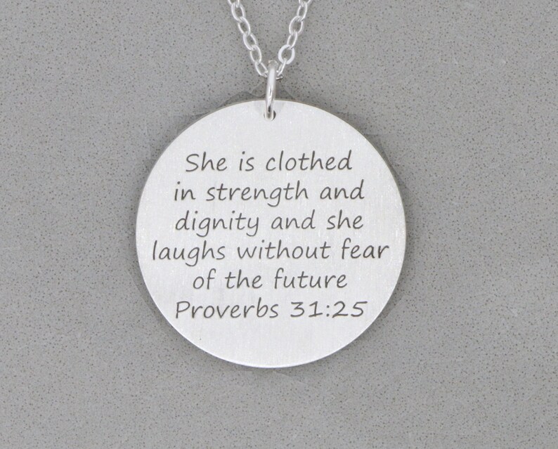 Silver necklace with large round pendant, engraved on front with Scripture verse in 6 lines of text is laying on gray stone background.