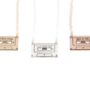 Three cassette tape charm necklaces in gold, silver, and rose gold lay on white background. Cassette tapes are engraved across top with our love songs, Ivy's Mix Tape, and Best of 1985. Charm hangs from either top corner to delicate chain.