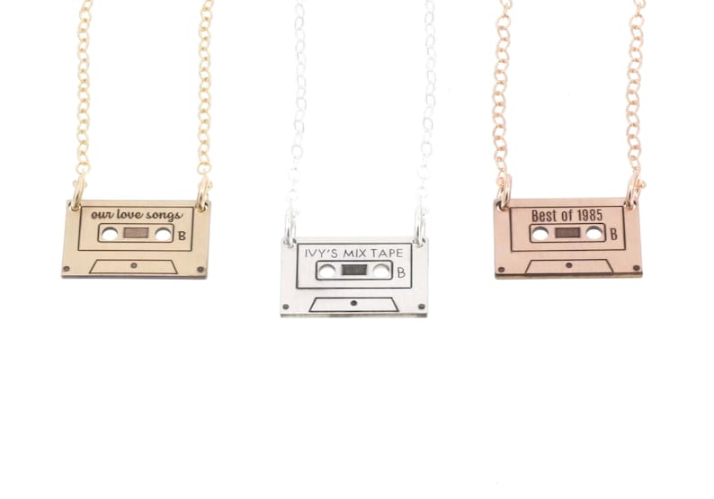 Three cassette tape charm necklaces in gold, silver, and rose gold lay on white background. Cassette tapes are engraved across top with our love songs, Ivy's Mix Tape, and Best of 1985. Charm hangs from either top corner to delicate chain.