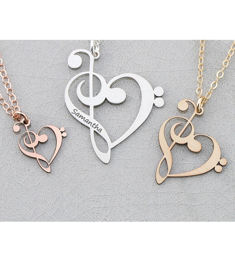Pode incluir: Tr&ecirc;s colares com pingentes em forma de cora&ccedil;&atilde;o em ouro rosa, prata e ouro. Cada colar apresenta um desenho de clave de sol dentro da forma de cora&ccedil;&atilde;o. O colar de prata tem o nome "Samantha" gravado. Os colares s&atilde;o exibidos em uma superf&iacute;cie cinza.