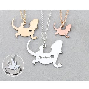 May include: Three silver necklaces with a bearded dragon silhouette pendant. The pendant on the left is gold, the middle pendant is silver with the name "Gordon" engraved on it, and the pendant on the right is rose gold. The necklaces are on a white background.