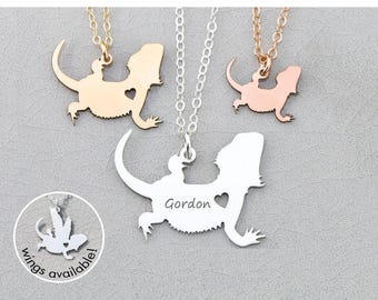 May include: Three silver necklaces with a bearded dragon silhouette pendant. The pendant on the left is gold, the middle pendant is silver with the name "Gordon" engraved on it, and the pendant on the right is rose gold. The necklaces are on a white background.