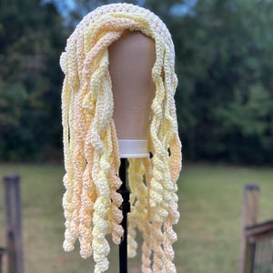 May include: A handmade crochet wig in shades of yellow and peach. The wig features a cap with long, spiraling strands that cascade down. The wig is displayed on a mannequin head against a blurred outdoor background.