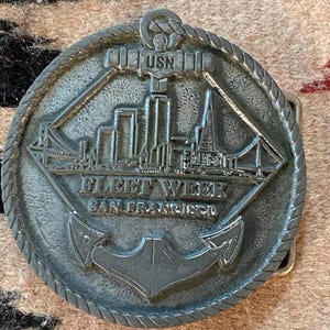May include: A silver-colored metal belt buckle featuring a detailed depiction of the San Francisco skyline, the Golden Gate Bridge, and an anchor. The text "FLEET WEEK SAN FRANCISCO" and "USN" are prominently displayed on the buckle.