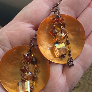 May include: A pair of copper-toned earrings. Each earring features a large, round, iridescent disc with dangling strands of amber and brown beads, crystals, and a square-cut crystal. The earrings have a decorative floral-like charm at the bottom.