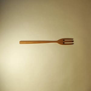May include: A wooden fork with three prongs. The fork is light brown in colour and is lying flat on a white surface.