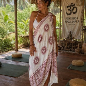 Può includere: Un pareo bianco con un motivo ripetuto di simboli solari color bordeaux. Il pareo è drappeggiato sulle spalle e intorno al corpo. Lo sfondo include uno studio di yoga con uno striscione che recita "ASHTANGA YOGA SHALA PALOLEM".