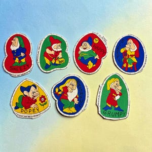 May include: Seven fabric patches featuring the Disney Seven Dwarfs: Sneezy, Doc, Happy, Sleepy, Dopey, Bashful, and Grumpy. Each patch is shaped with a white border and includes the character's name printed on the fabric, with instructions to leave open for stuffing.
