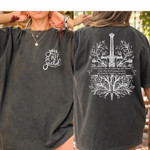 Może przedstawiać: Ciemnoszary t-shirt z krótkim rękawem z białym nadrukiem. Z przodu znajduje się napis "you do not yield" wykonany pismem odręcznym. Z tyłu widnieje duża ilustracja miecza otoczonego misternymi kwiatowymi pnączami.