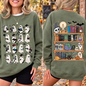 May include: An olive green crewneck sweatshirt featuring a graphic print on the front and back. The front displays multiple white ghosts reading books. The back shows a wooden bookshelf filled with books, a skull, a candle, and a cauldron.