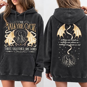 Puede incluir: Una sudadera con capucha gris carbón que presenta un gráfico frontal con el texto The Valkyrie Cycle y Three Valkyries, One Bond, junto a ilustraciones de montañas y alas. La parte posterior muestra alas, montañas y el texto nothing can break us.