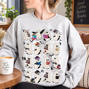May include: A light gray crewneck sweatshirt featuring a repeating pattern of white ghosts reading various books. The ghosts are depicted in multiple poses, some with bats, stars, and small icons. The design is printed on the front of the garment.