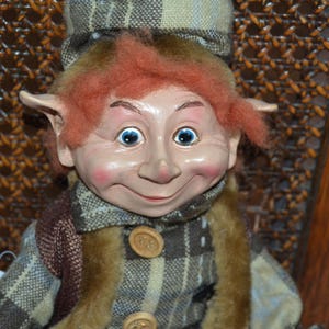 Peut inclure: Une poupée fantaisiste avec un visage souriant, des yeux bleus et des cheveux roux. La poupée porte un chapeau, un manteau et une écharpe à carreaux avec une garniture en fausse fourrure. De gros boutons en bois ornent le manteau.