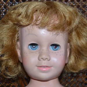 May include: A vintage doll with a fair complexion, blue eyes, and freckles. The doll has a soft pink mouth and light brown, curly hair. The doll's face is the focal point, with a close-up view of its features.