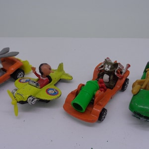 May include: Five toy cars featuring cartoon characters. A yellow and green airplane with a red-headed pilot, a green car with a brown dog, an orange car with a green cannon, an orange car with a gray cat, and an orange car with a gray rabbit.