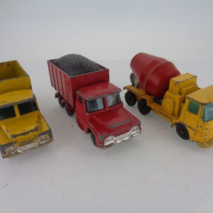 Puede incluir: Tres camiones de juguete, un camión volquete amarillo, un camión rojo con una carga negra y un camión mezclador de cemento amarillo.
